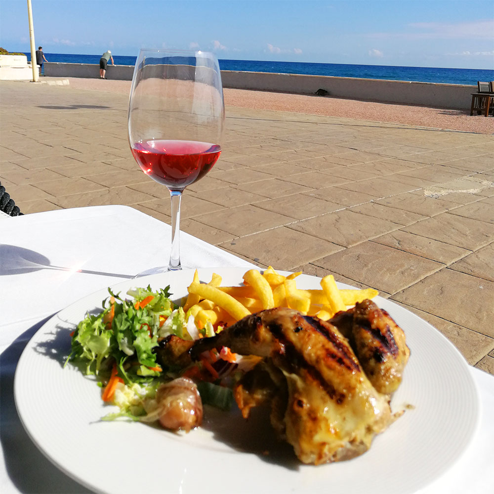 Tallrik med kyckling och pommes frites, glas rödvin, utsikt över havet.