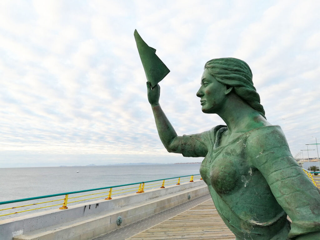 Staty av kvinna som står och vinkar mot havet.