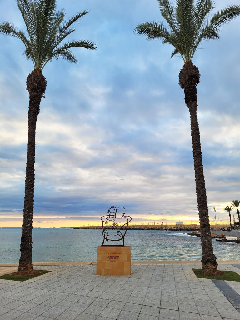 Skulptur bredvid två palmer vid havet.