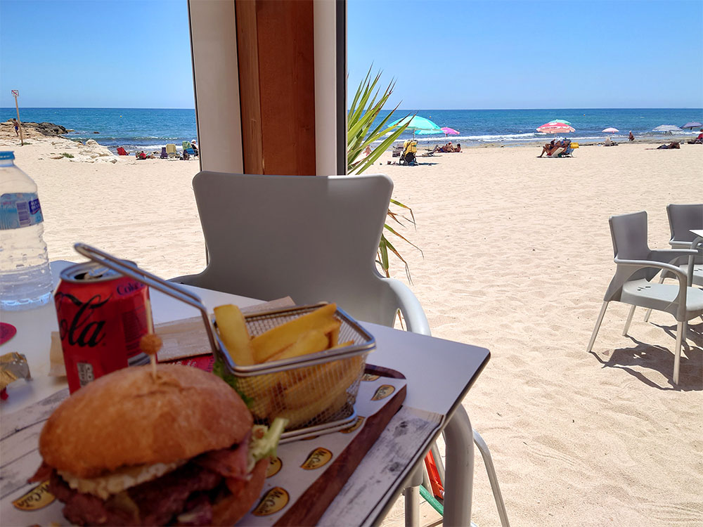 Hamburgare, pommes och coca cola på en servering mitt på sandstranden.